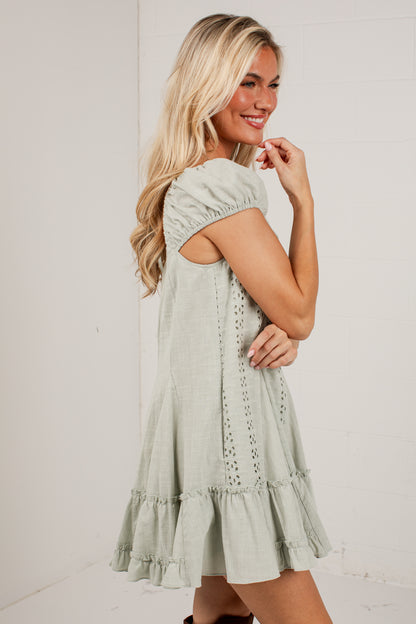 A woman modeling a light sage green eyelet mini dress with short puff sleeves and a tiered ruffle hem, photographed against a white studio wall.