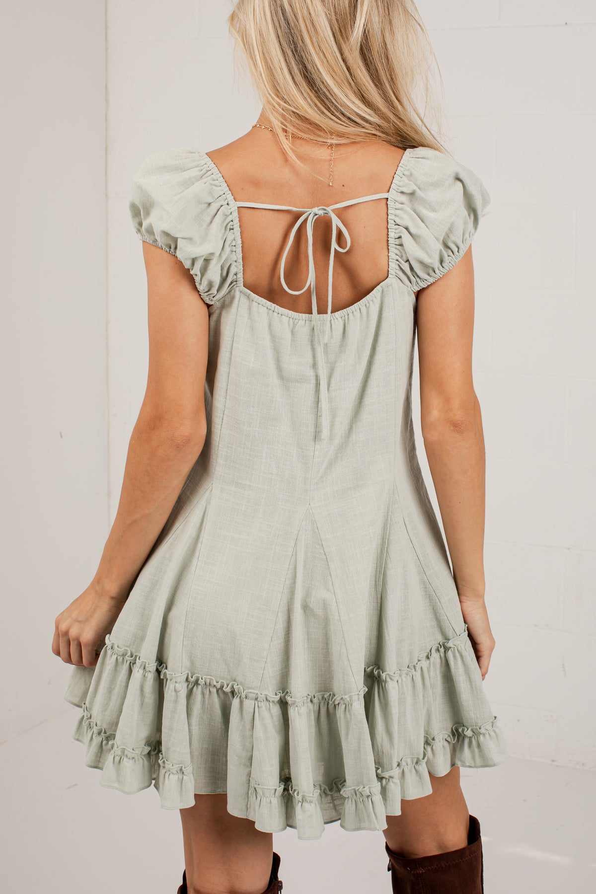 A woman wearing a sage green linen-look mini dress with puff sleeves, a tie-back neckline, and a ruffled hem, shown from the back against a white wall.