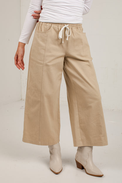 A person wearing beige wide-leg cropped pants with a white drawstring waist, paired with a white long-sleeve top and light beige ankle boots, photographed against a plain light wall.