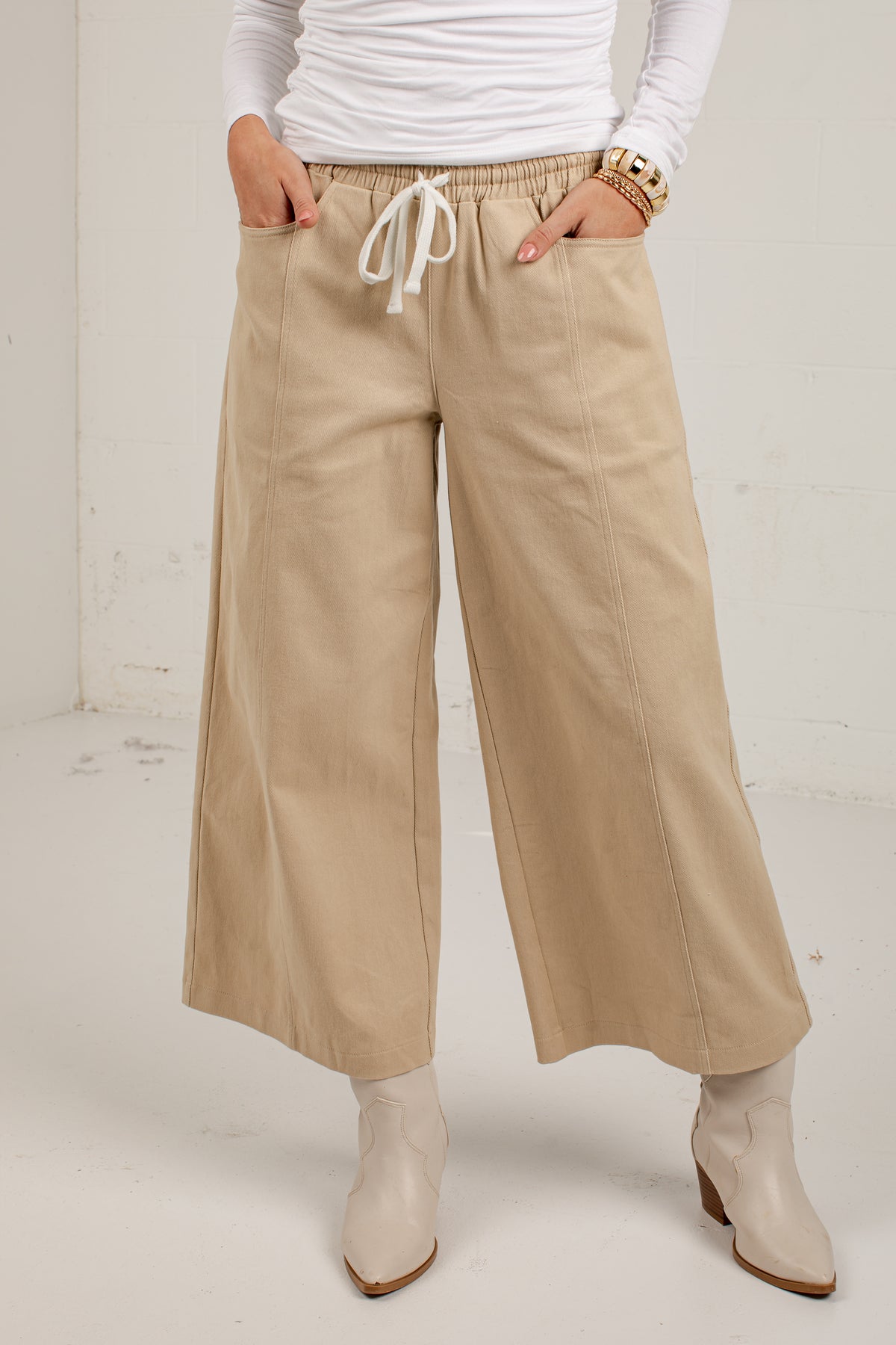 A person wearing beige wide-leg cropped pants with an elastic drawstring waist, paired with a white long-sleeve top and white ankle boots, standing against a plain off-white wall.
