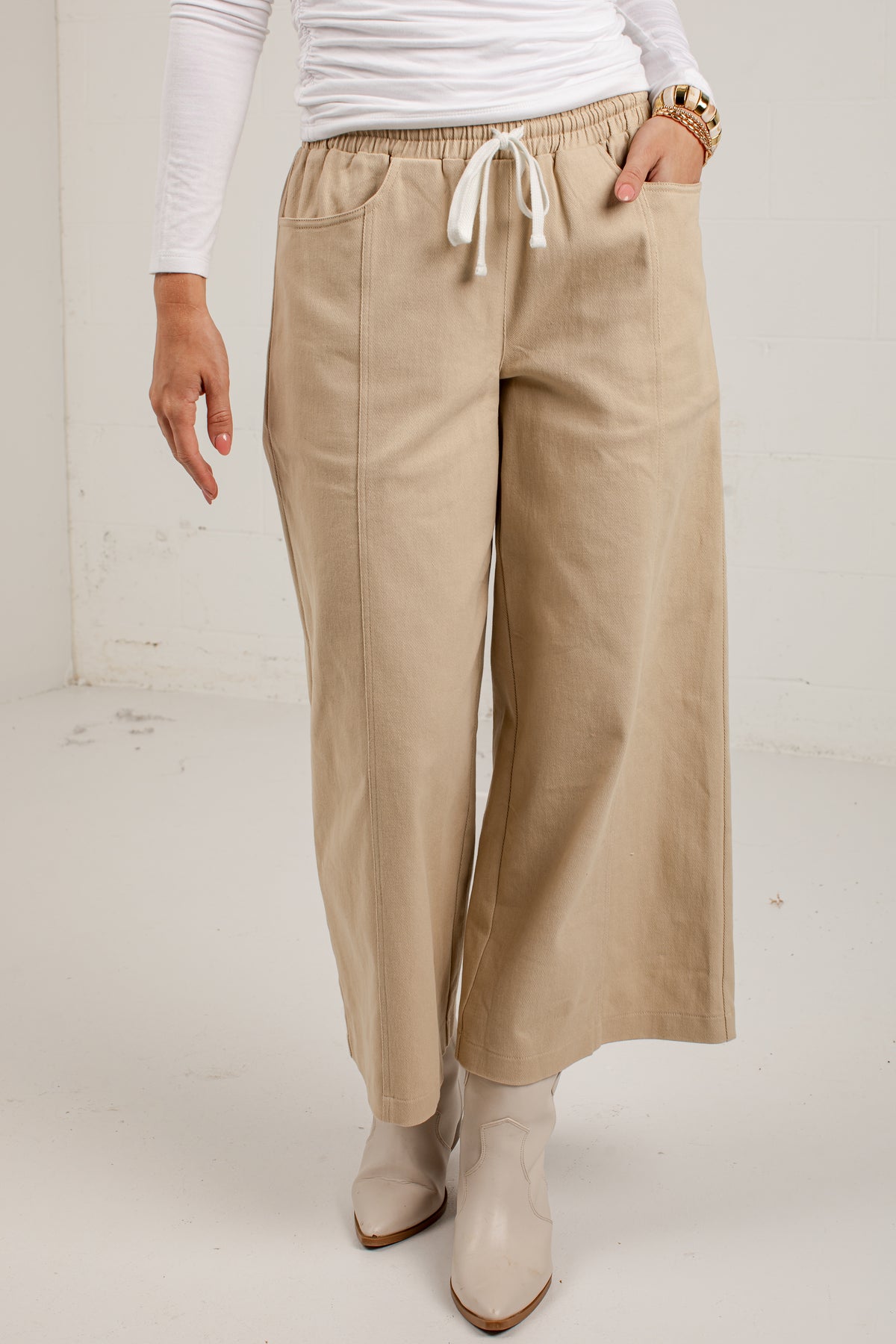 A person wearing beige wide-leg cropped pants with an elastic waistband and white drawstring, paired with a white long-sleeve top and beige ankle boots.