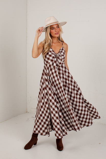 A woman modeling a brown and white gingham maxi dress with a tie-front detail, paired with a cream wide-brim hat and brown ankle boots, standing in a white studio corner.