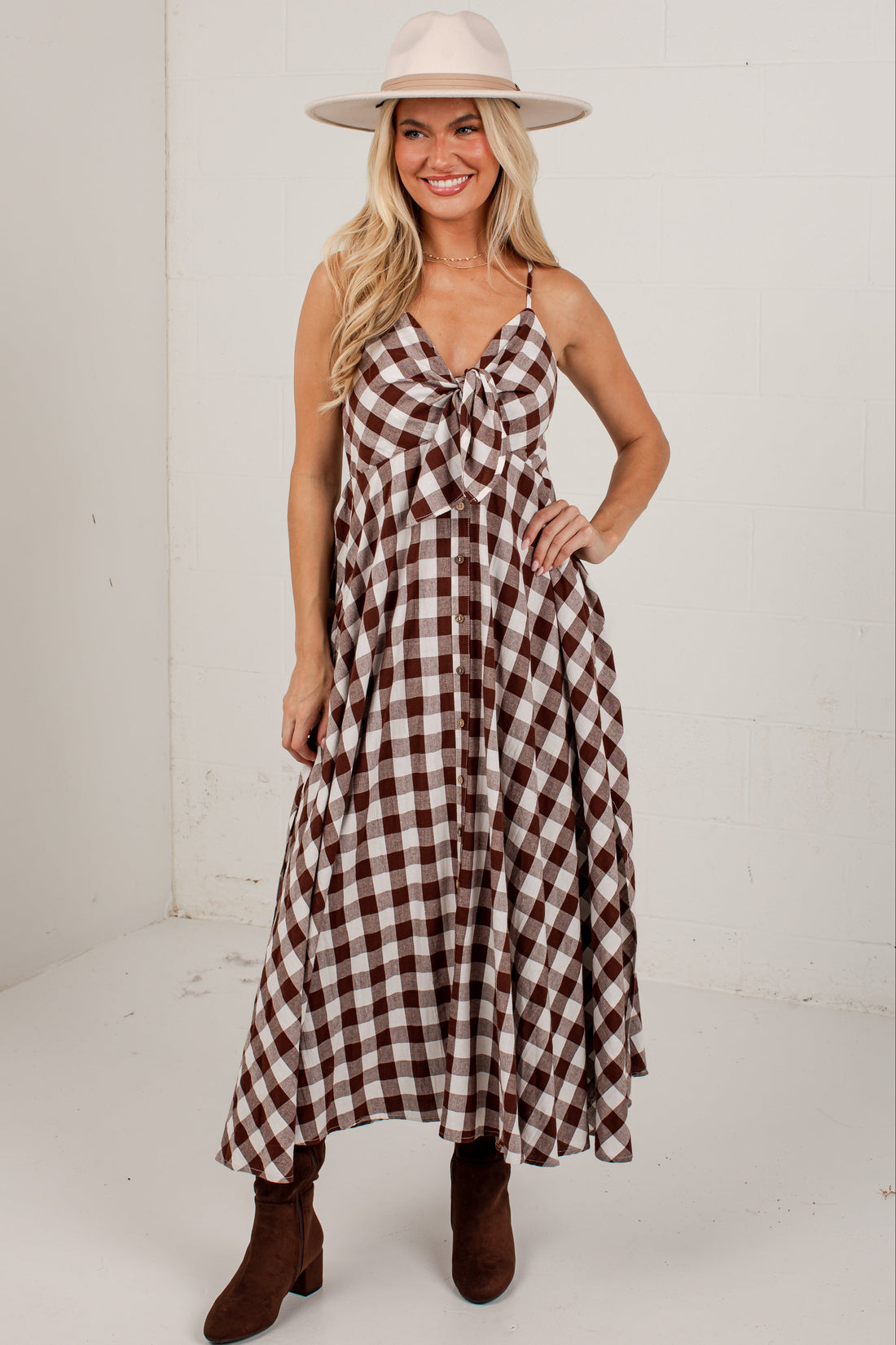 A woman modeling a brown and beige gingham maxi dress with spaghetti straps and a front tie detail, paired with a beige wide-brim hat and brown ankle boots, in a white studio setting.