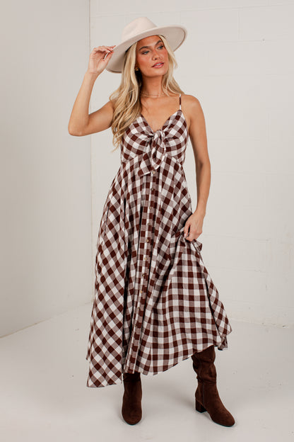 A woman modeling a brown and white gingham maxi dress with spaghetti straps and a knotted bodice, styled with a wide-brimmed cream hat and brown knee-high boots against a white studio background.