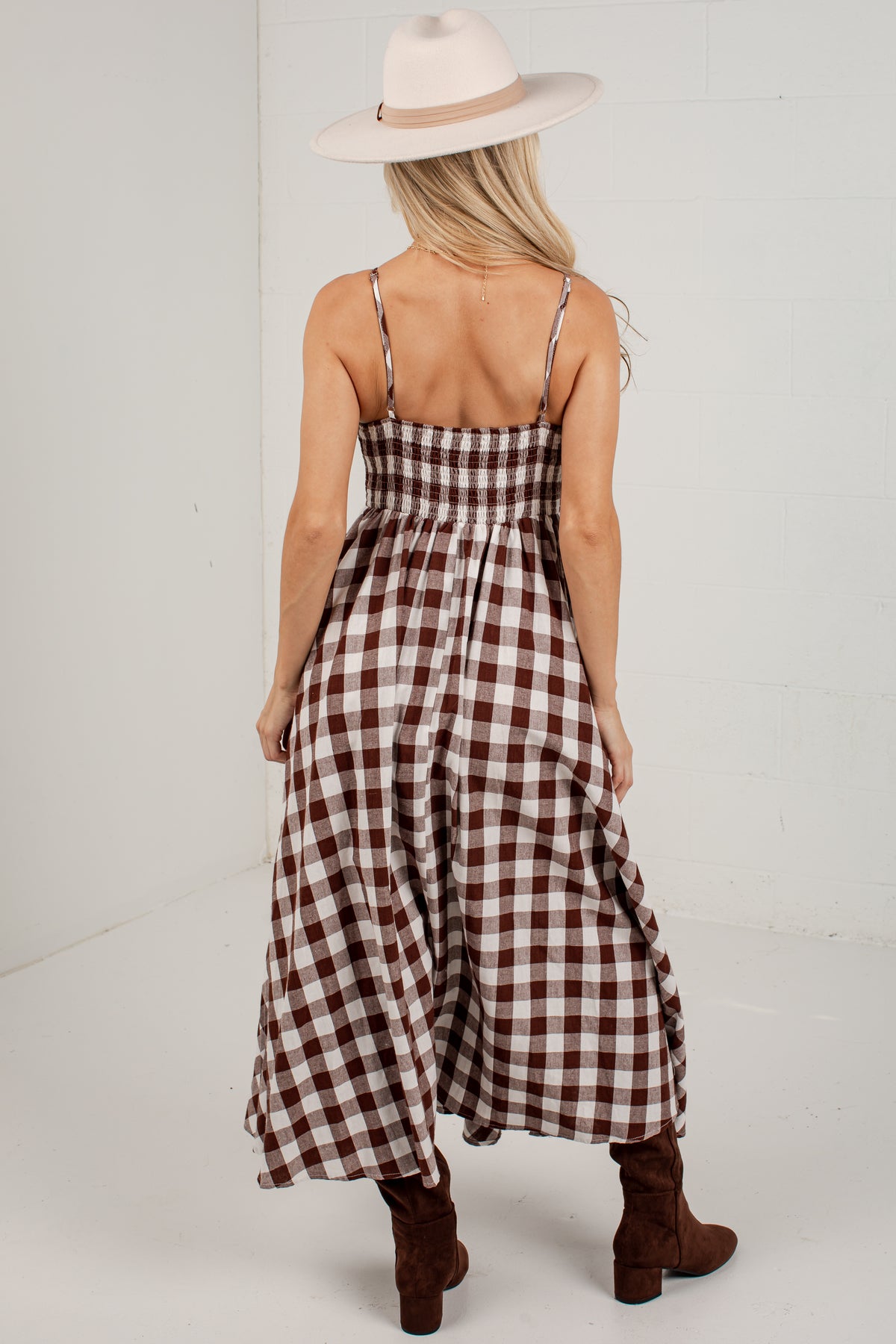 A woman shown from the back wearing a brown and cream gingham maxi dress with thin spaghetti straps, a smocked bodice, and a flared skirt. She is also wearing a cream wide-brim hat and brown suede ankle boots, standing against a white studio background.