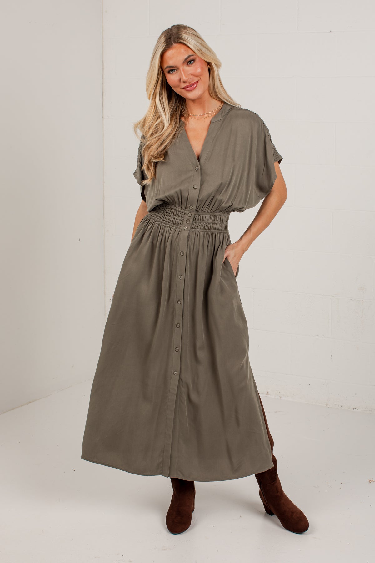 A woman modeling a loose, short-sleeve midi dress in a muted olive-gray or taupe-brown tone, paired with brown ankle boots, against a plain white studio wall.