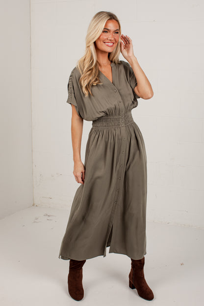 A woman modeling a short-sleeve, button-front midi dress in a muted olive green color, paired with brown suede boots, standing against a white studio wall.