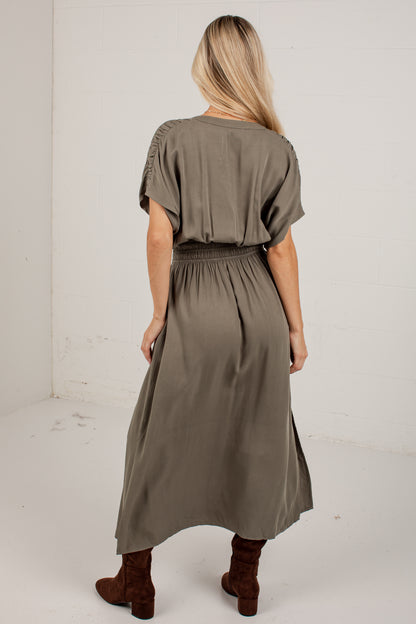 A woman viewed from behind wearing an olive green midi dress with short flutter sleeves, a gathered waist, and brown ankle boots, standing against a plain white wall.