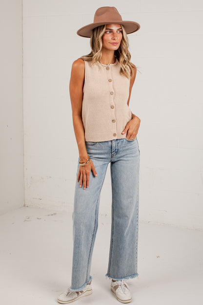 A woman modeling a sleeveless beige knit button-front top with light-wash wide-leg jeans, a brown wide-brim hat, and white sneakers, standing against a plain white wall.