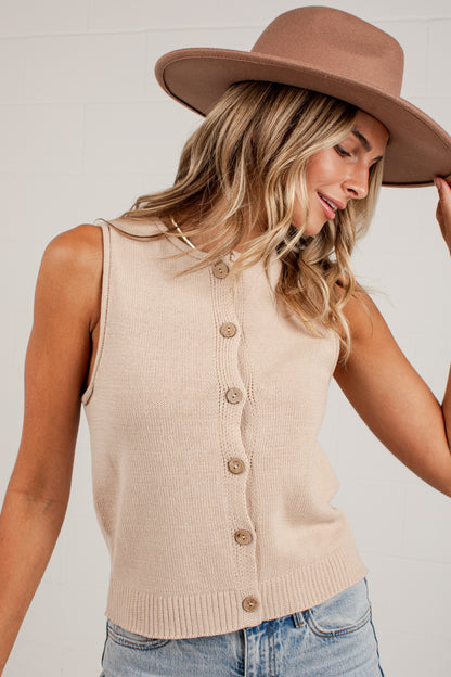 A woman wearing a beige sleeveless ribbed knit cardigan vest with brown buttons, paired with a brown wide-brim hat and light blue jeans, against a white brick wall background.
