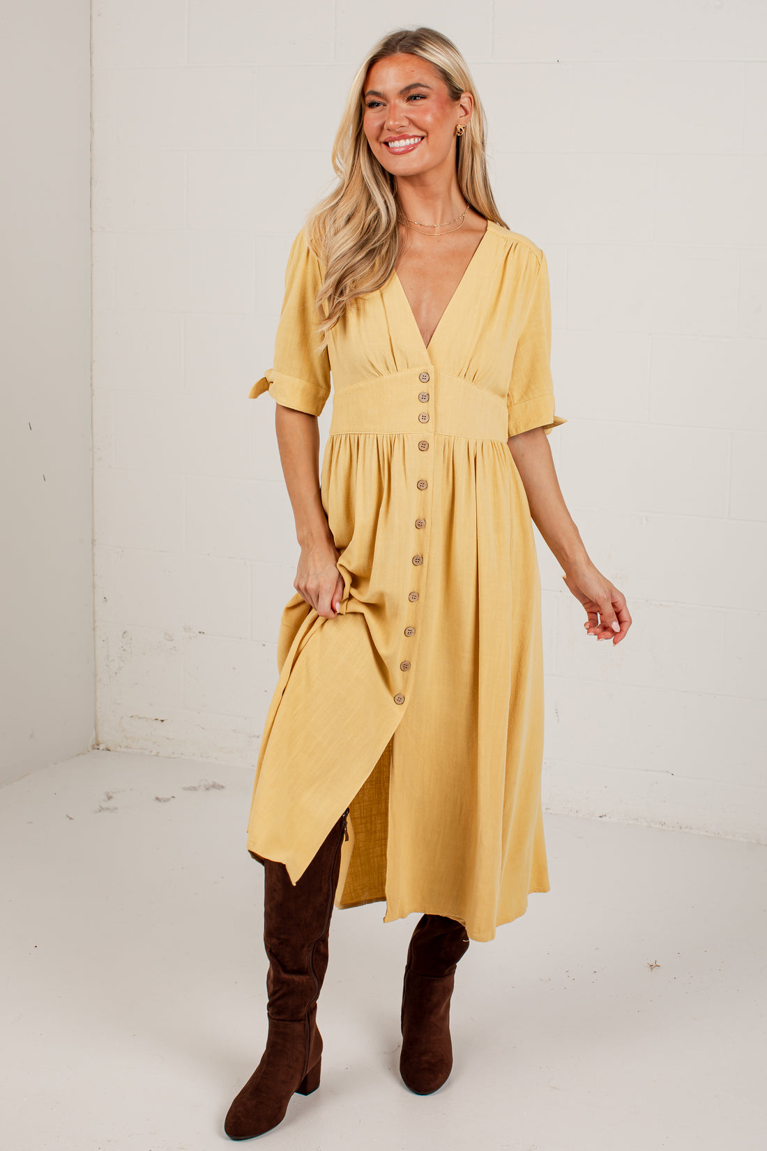 A woman modeling a mustard yellow midi dress with a button-down front, short sleeves, and a gathered waist, paired with brown knee-high boots against a plain white studio background.