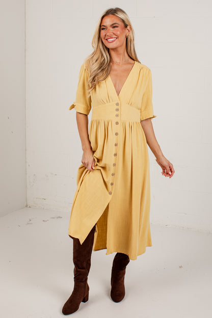 A woman modeling a mustard yellow midi dress with a button-down front, short sleeves, and a gathered waist, paired with brown knee-high boots against a plain white studio background.