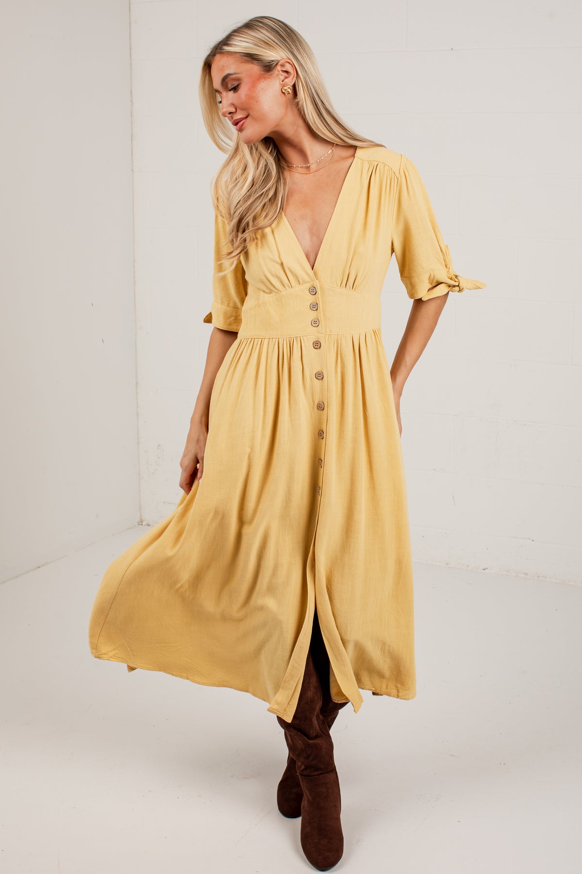 A woman modeling a mustard yellow midi dress with puff sleeves, a V-neckline, and a button-down front, paired with brown boots in a bright studio setting.