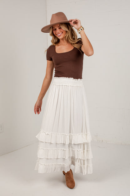 A woman modeling a bohemian-style outfit consisting of a brown short-sleeve scoop-neck top, a long white tiered ruffle maxi skirt, a brown wide-brim hat, and brown ankle boots, standing against a white brick wall.