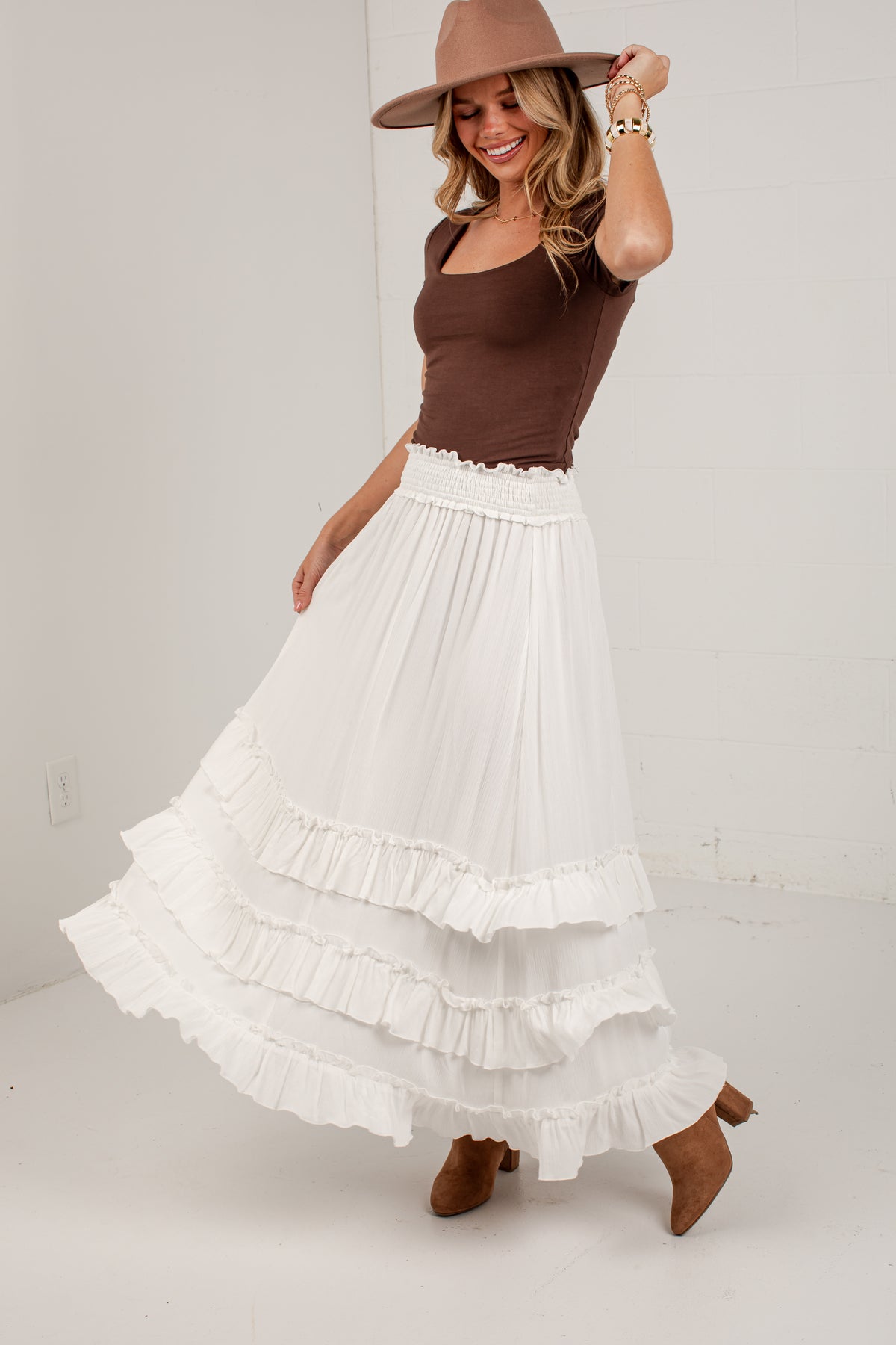 A woman modeling a long white tiered maxi skirt with a ruffled waistband, paired with a brown scoop-neck top, brown hat, and brown boots, standing against a white wall.
