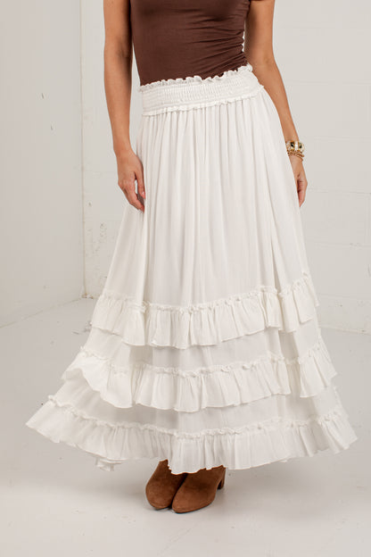 A model wearing a long white tiered maxi skirt with a smocked waistband, paired with a brown top and brown suede boots.