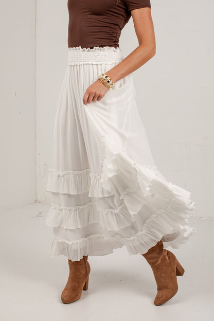 A white tiered maxi skirt with a ruffled hem, shown on a model wearing a brown top and brown suede ankle boots against a white studio background.