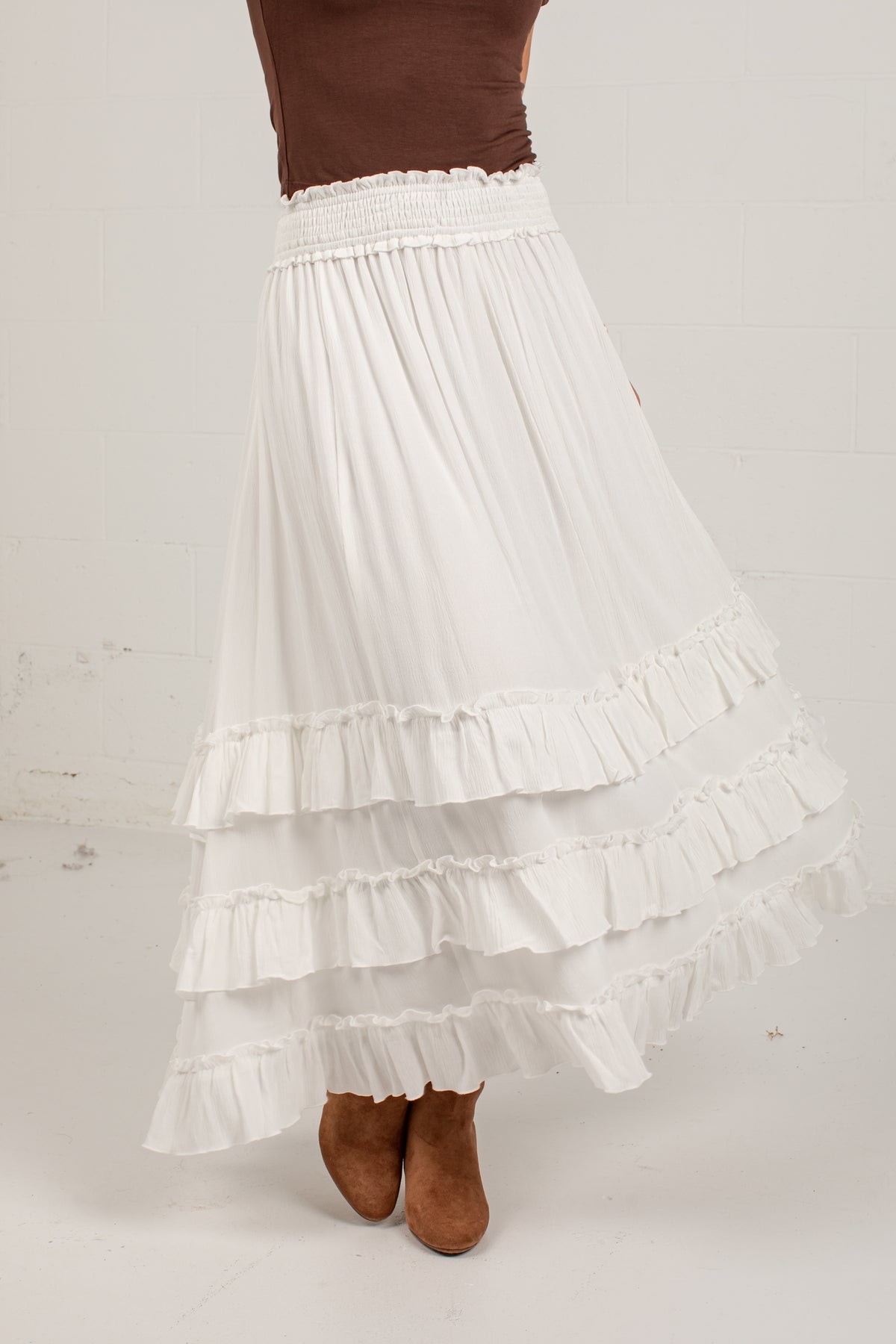 A white tiered ruffle maxi skirt with a smocked elastic waistband, shown on a model wearing brown suede boots against a light off-white background.