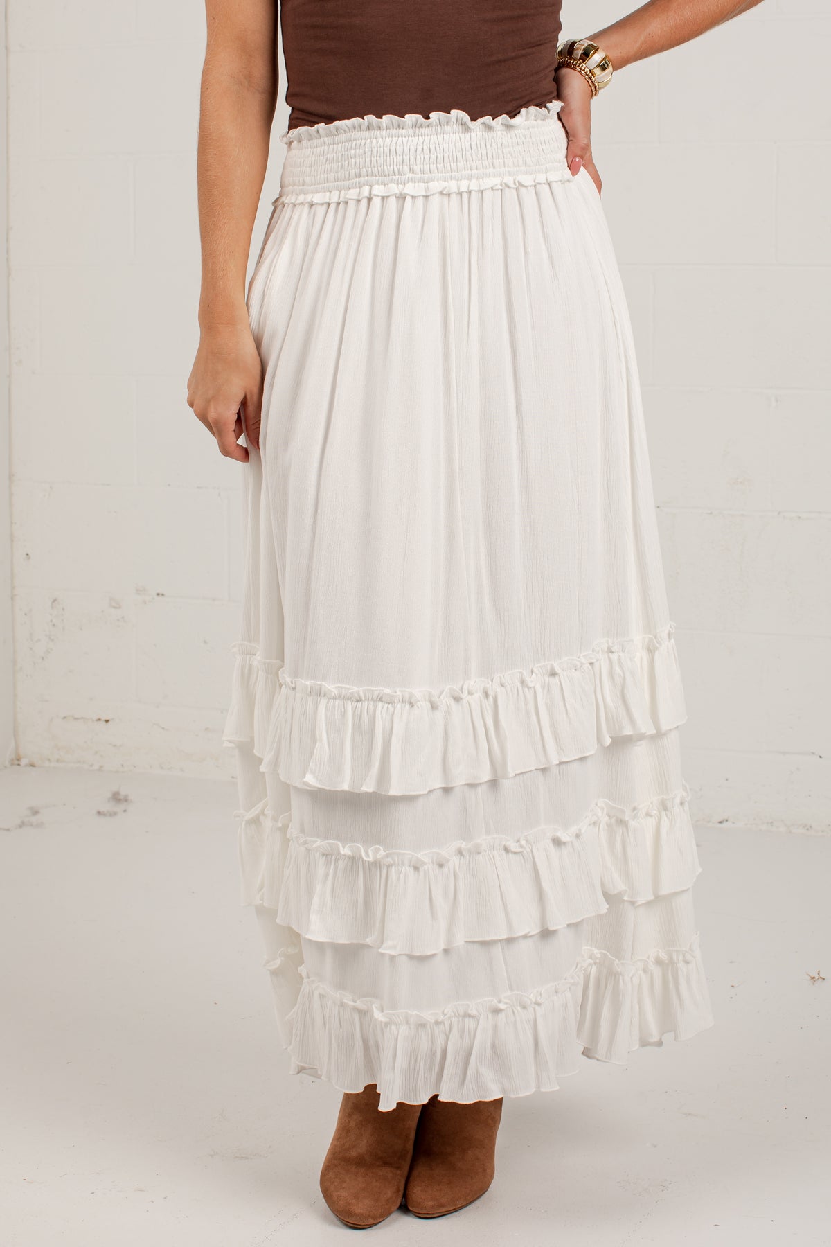 A white tiered maxi skirt with a smocked elastic waistband, shown on a model wearing brown suede boots and a brown top against a white brick wall.