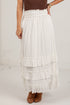 A white tiered maxi skirt with a smocked elastic waistband, shown on a model wearing brown suede boots and a brown top against a white brick wall.