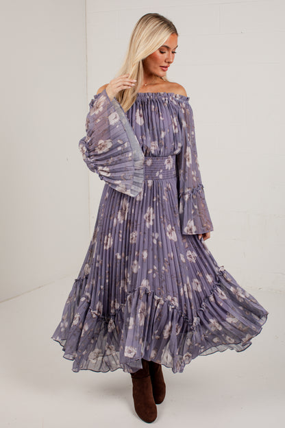 A woman modeling an off-the-shoulder, long-sleeved maxi dress with a purple and white floral print, paired with brown boots, in a bright studio setting.