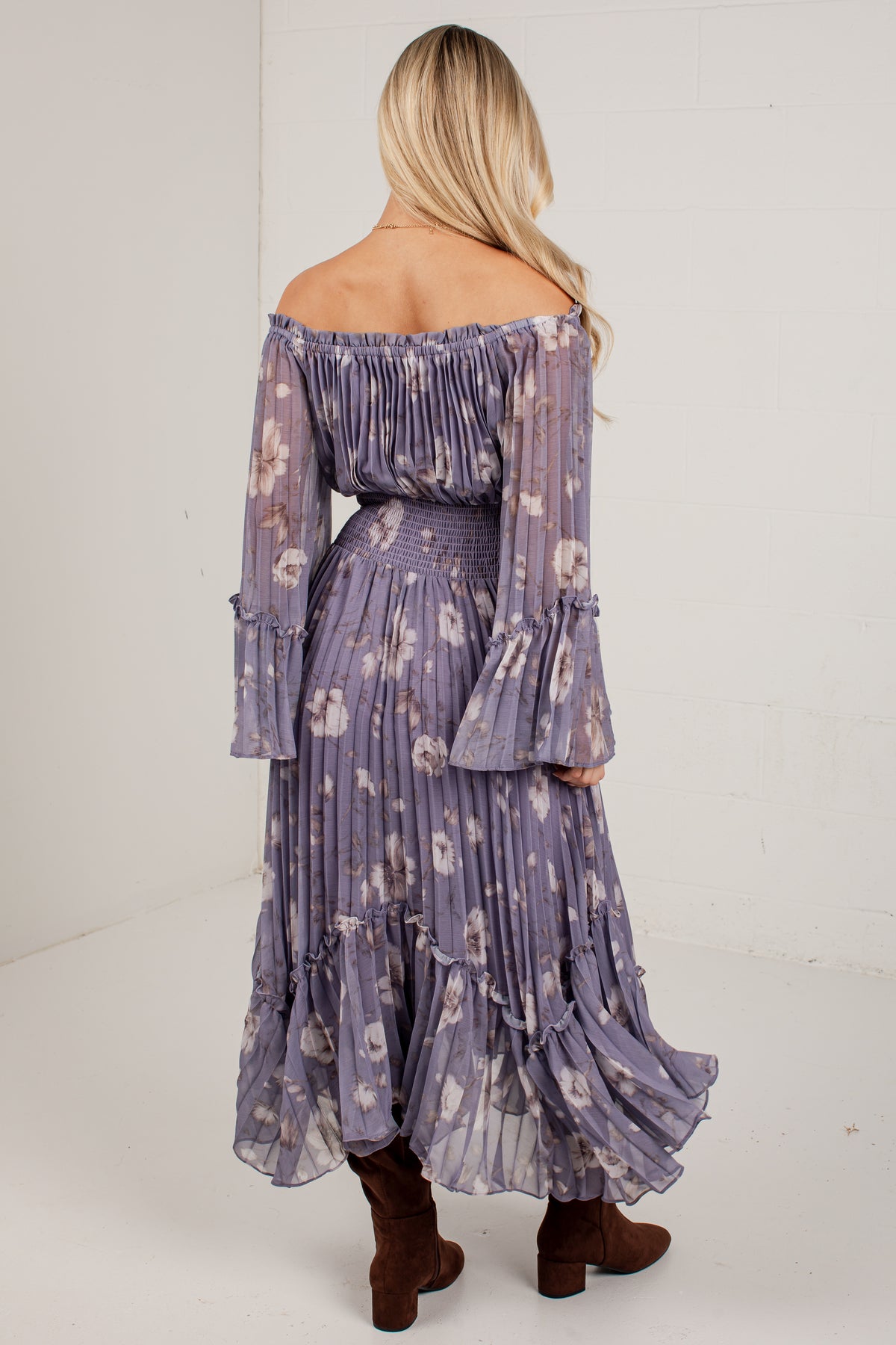 A woman shown from the back wearing a lavender off-the-shoulder maxi dress with a floral print, pleated long sleeves, and a ruffled tiered hem, paired with brown ankle boots against a white studio background.