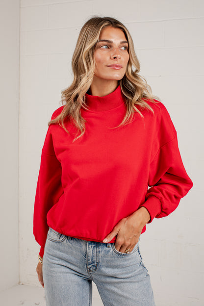 A woman wearing a red mockneck sweatshirt and light blue jeans, standing against a solid light gray background.