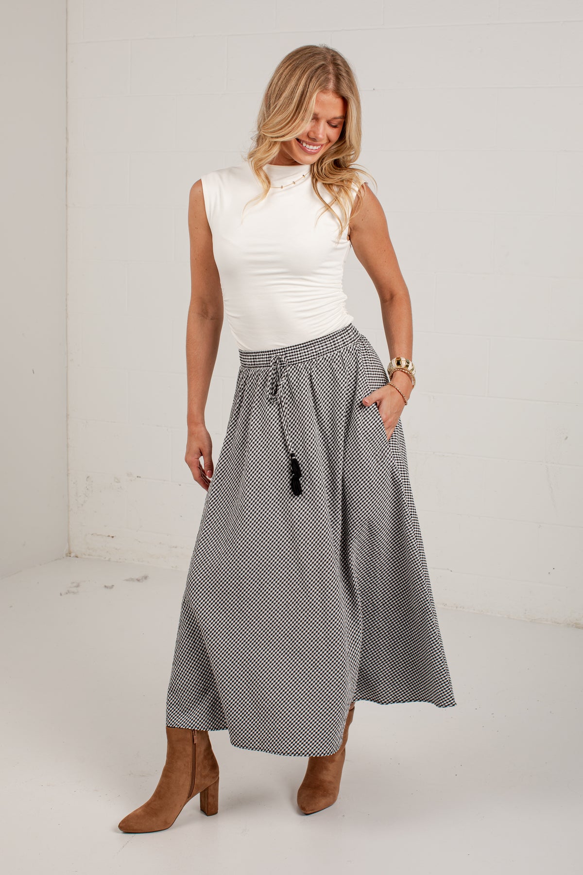 A woman modeling a sleeveless white mock-neck top and a high-waisted, knee-length gray and white micro-check skirt with a drawstring waist and pockets, paired with brown ankle boots.