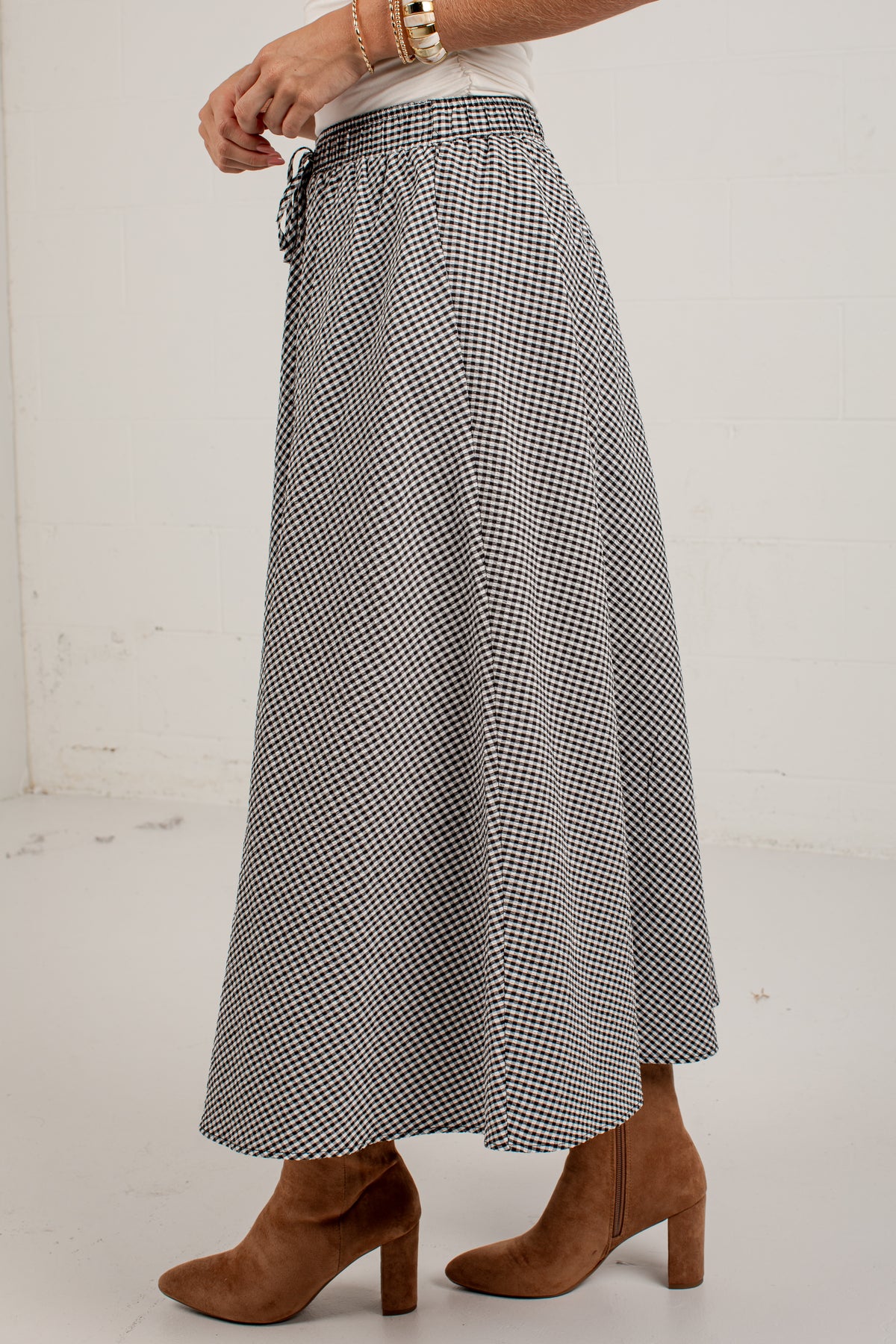 A person wearing a black and white gingham patterned maxi skirt with a drawstring waist, styled with a white top and brown suede block-heel ankle boots.