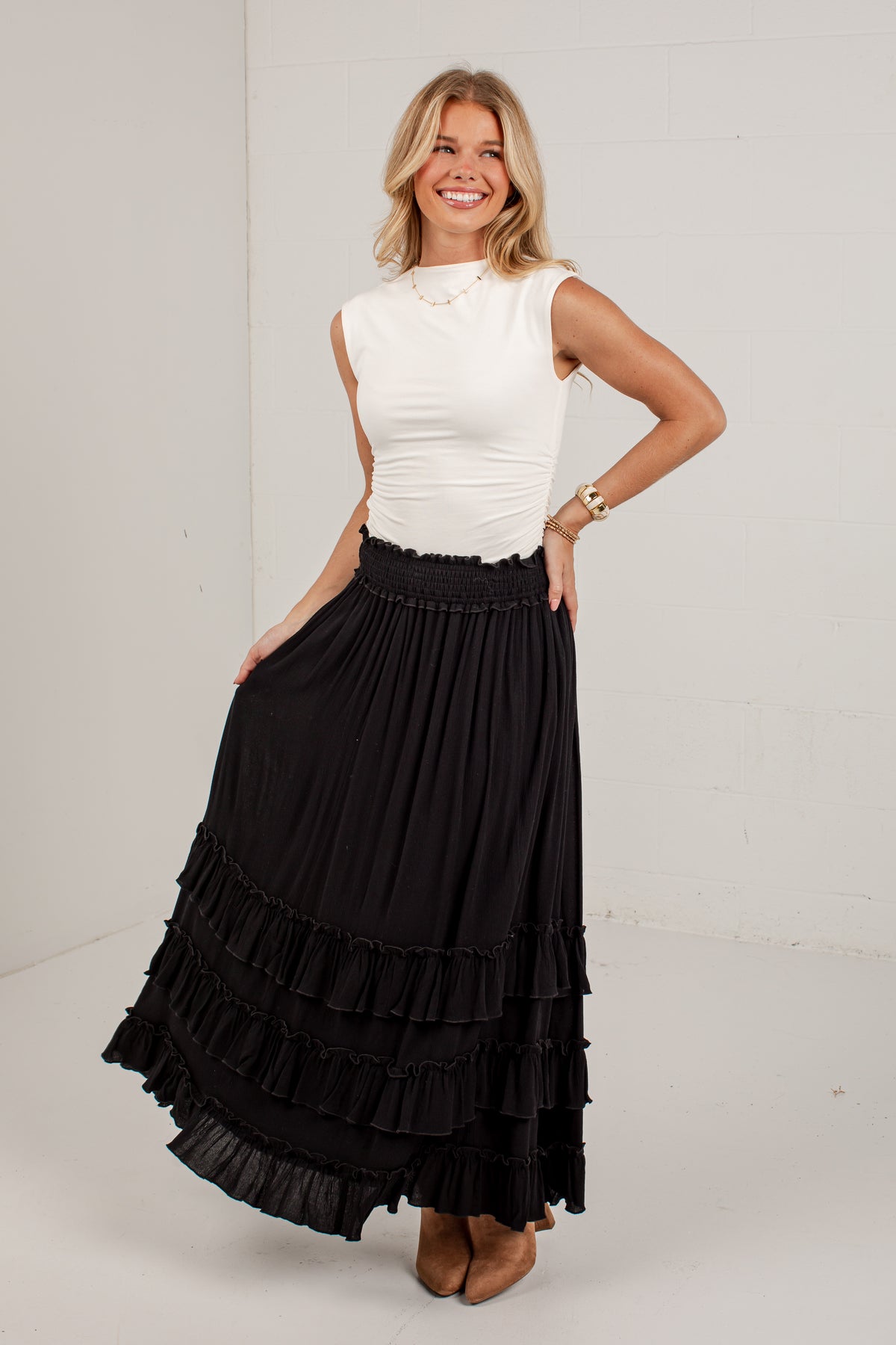 A woman modeling a long black tiered maxi skirt paired with a white sleeveless top, standing against a white studio background.