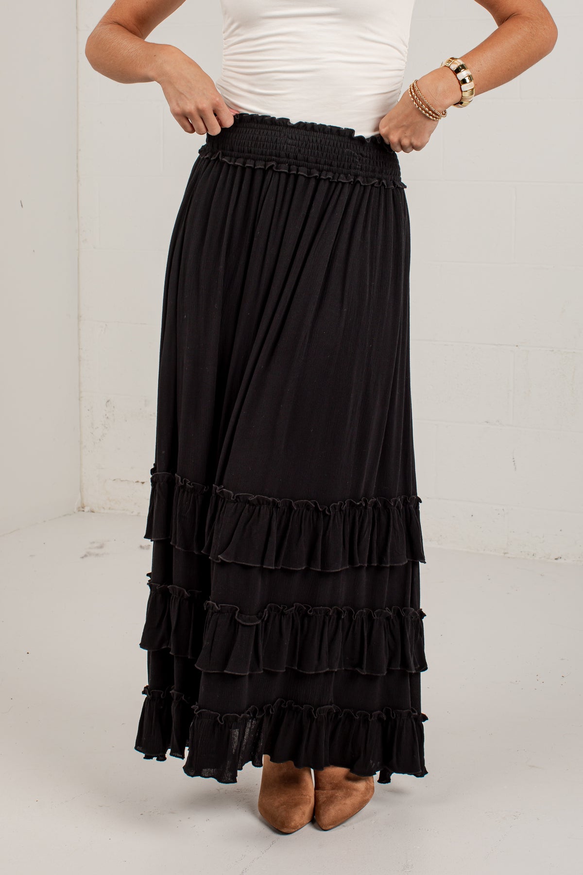 A black tiered maxi skirt with a smocked elastic waistband, shown on a model wearing a white top and brown shoes against a white studio background.