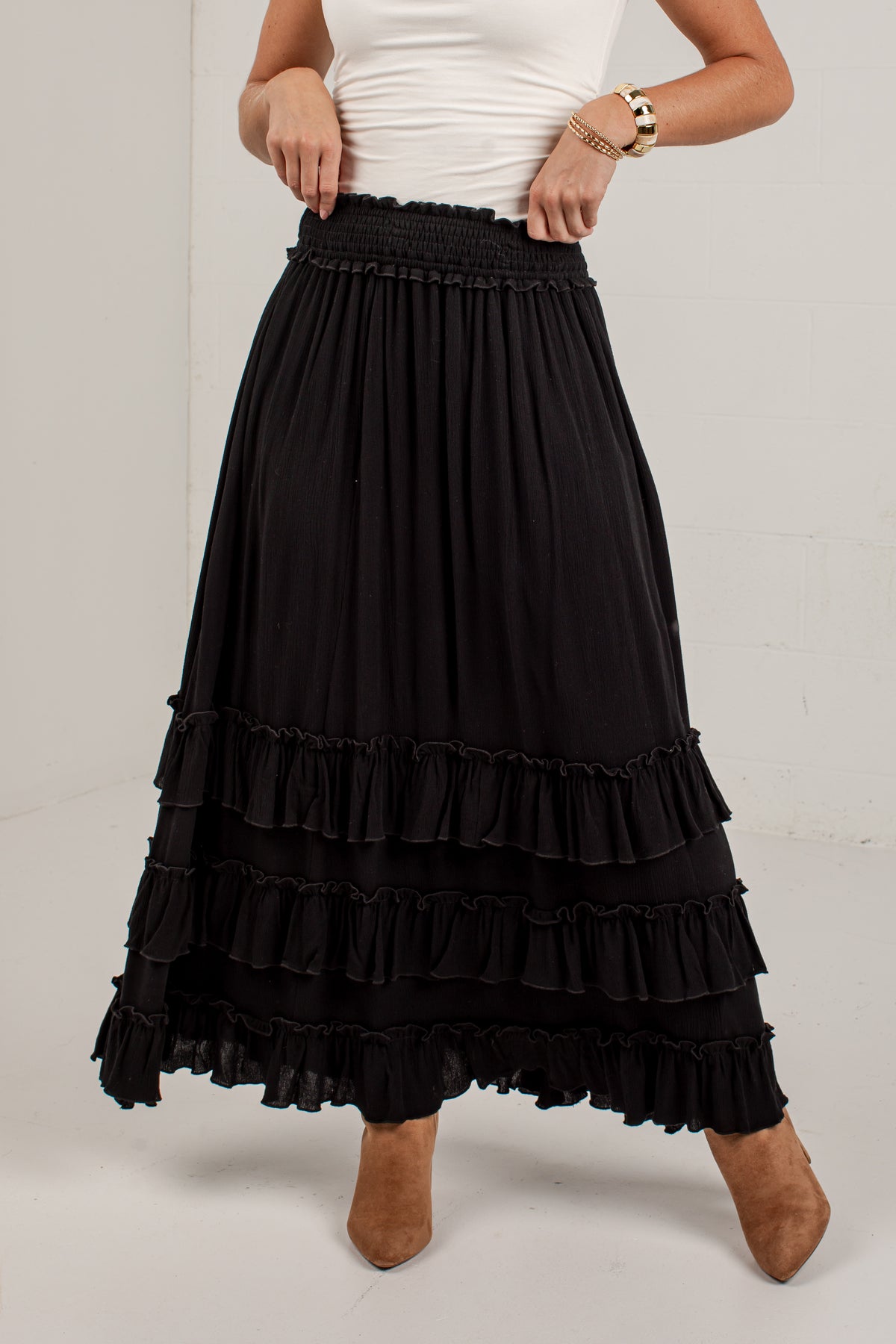 A black tiered maxi skirt with a smocked elastic waistband, worn by a model with a white top and brown boots against a white studio background.
