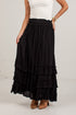 A black tiered maxi skirt with an elastic waistband, worn by a model with a white top and brown suede boots against a white brick wall.