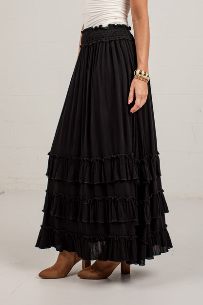 A black tiered maxi skirt with a smocked elastic waistband, shown on a model wearing a white top and brown boots against a white studio wall.