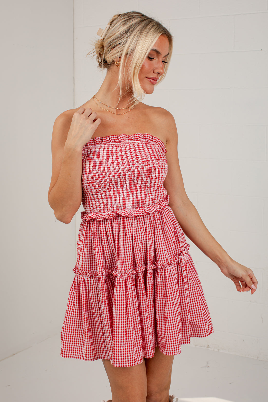 A woman modeling a red and white gingham strapless mini dress with a smocked bodice and tiered skirt, standing against a white brick wall.