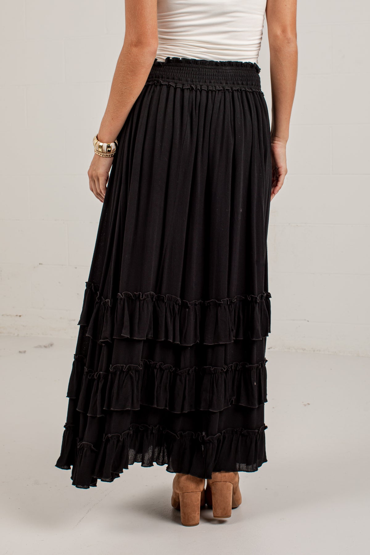 A black tiered maxi skirt with a smocked waistband, shown on a model from the back wearing a white top and brown block-heel sandals.