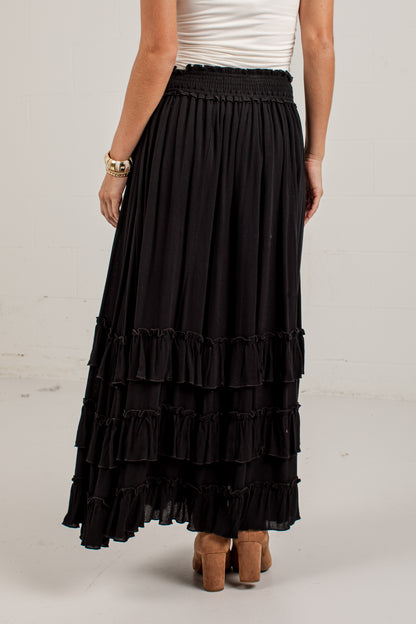 A black tiered maxi skirt with a smocked waistband, shown on a model from the back wearing a white top and brown block-heel sandals.