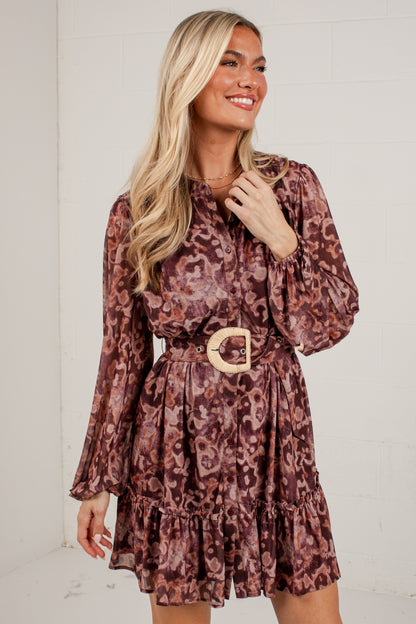 A woman modeling a long-sleeve mini dress with a brown and dusty rose floral pattern, featuring a matching belt with a large gold buckle.