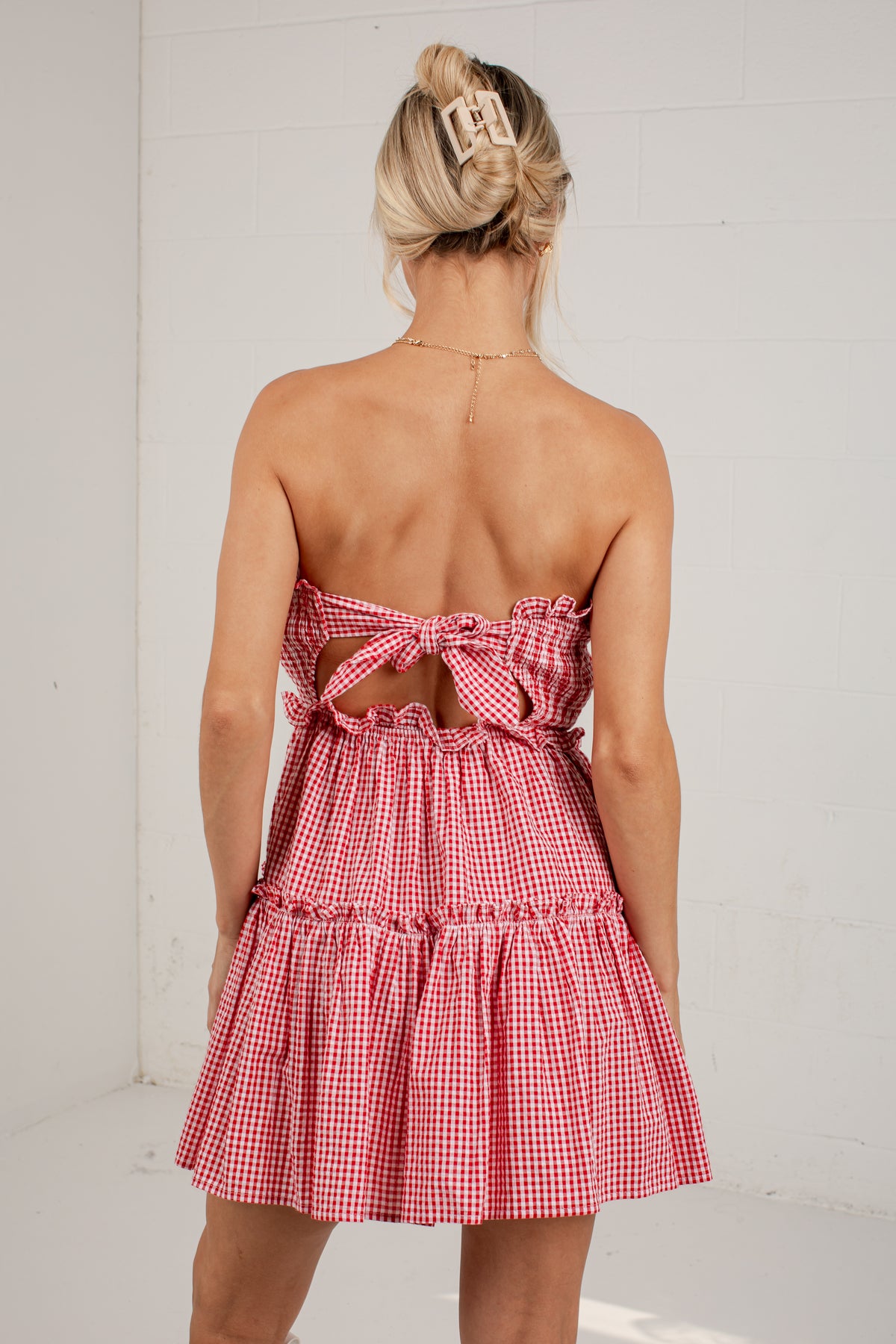 A woman shown from the back wearing a strapless red and white gingham mini dress with a bow detail at the back, standing against a white wall.