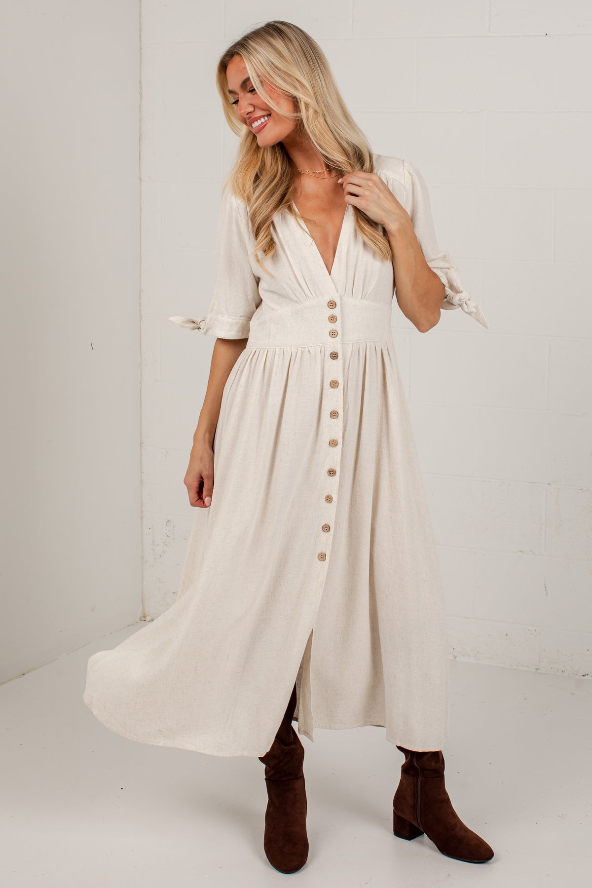 A woman modeling a cream-colored midi dress with a button-down front, tie details on the sleeves, and brown suede ankle boots, standing against a white brick wall.