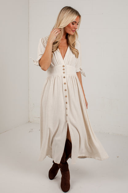 A woman modeling a cream-colored midi dress with a button-down front, ruffled sleeves, and a high slit, paired with brown boots in a bright studio setting.
