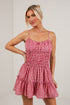 A woman modeling a red and white gingham mini dress with thin spaghetti straps, a smocked bodice, and a tiered ruffle skirt, set against a white brick wall.