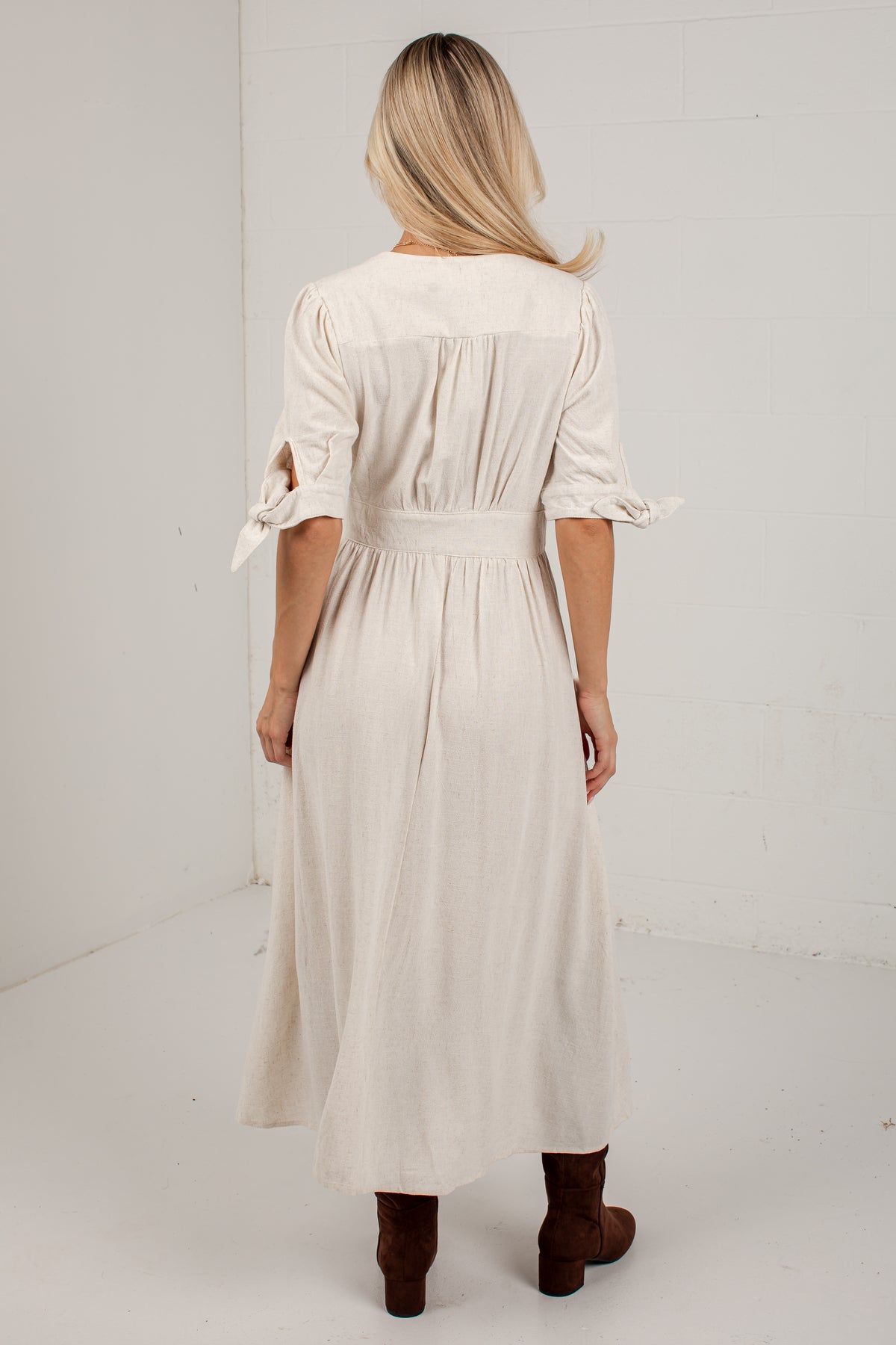A woman viewed from behind wearing a cream-colored midi dress with three-quarter sleeves, tie details at the cuffs, and a gathered waist, paired with brown ankle boots in a white studio setting.