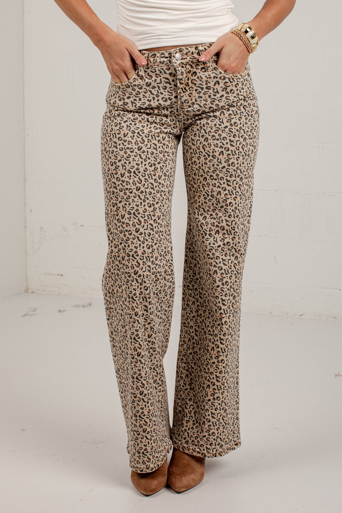 A person wearing leopard-print pants with a white top and brown shoes, standing against a plain white wall.