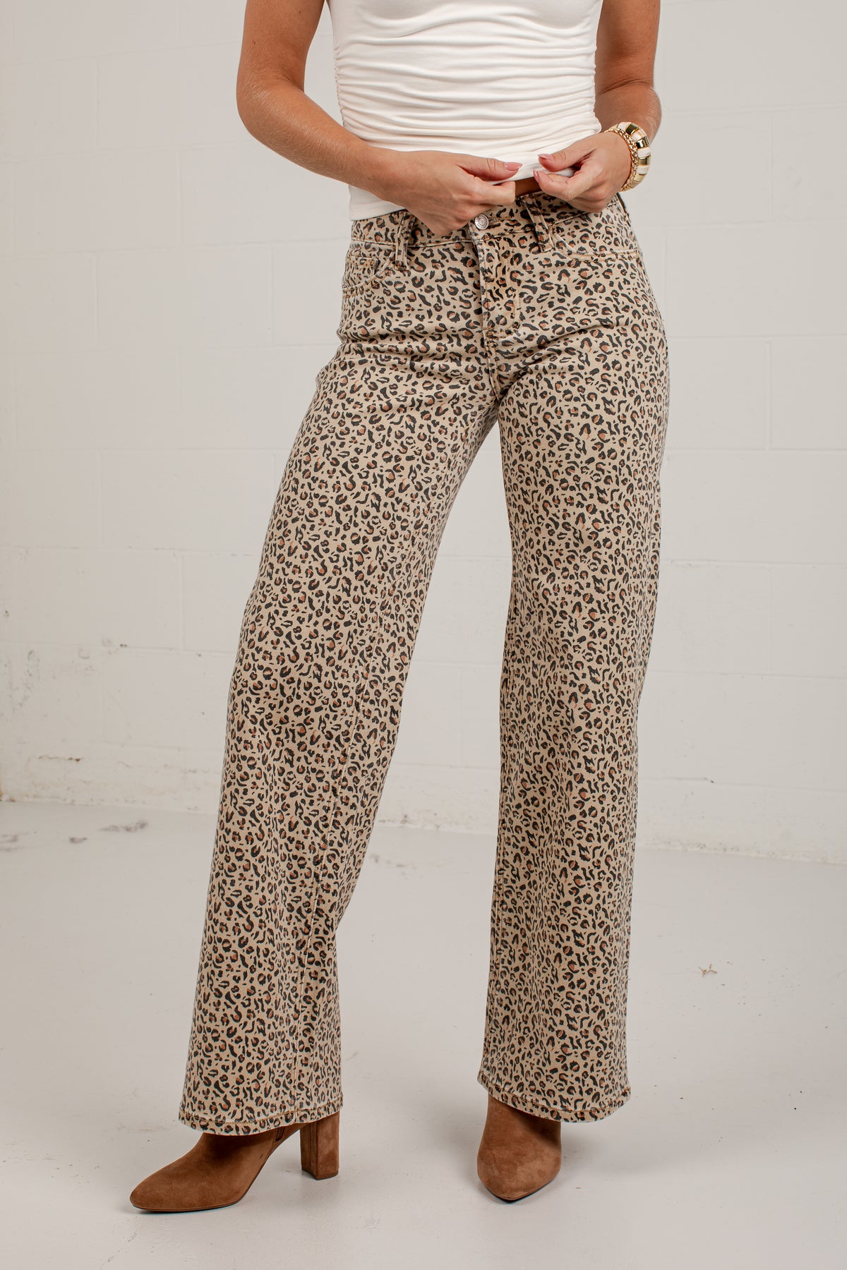 A person wearing leopard-print wide-leg pants with a white ribbed top and brown heeled boots, standing against a plain off-white wall.