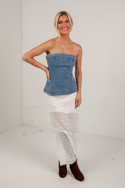 A woman modeling a strapless denim corset-style top paired with a white textured skirt and brown ankle boots, standing against a white studio wall.