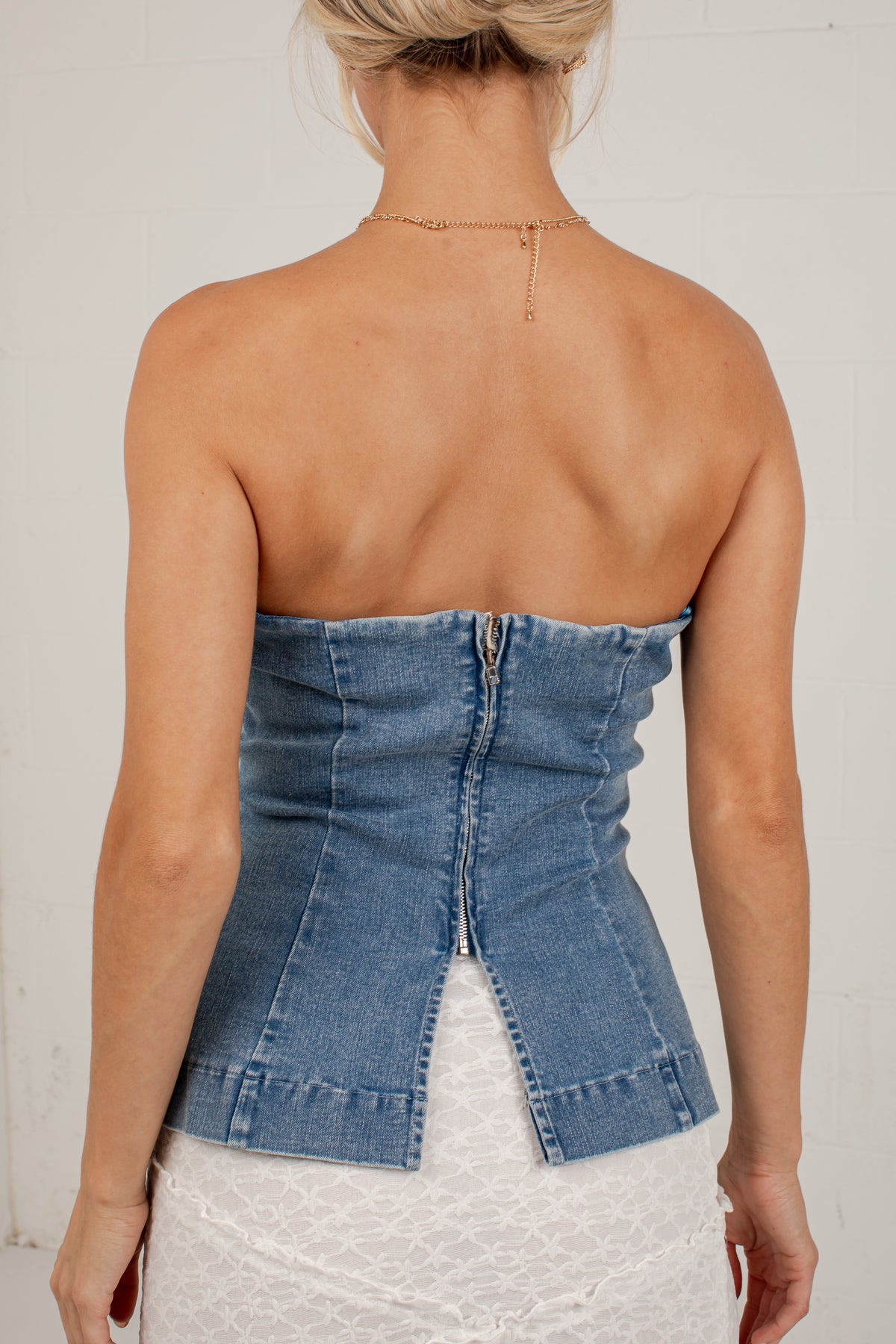 Rear view of a woman wearing a strapless blue denim corset-style top with a center back zipper, paired with a white textured skirt, against a plain light background.