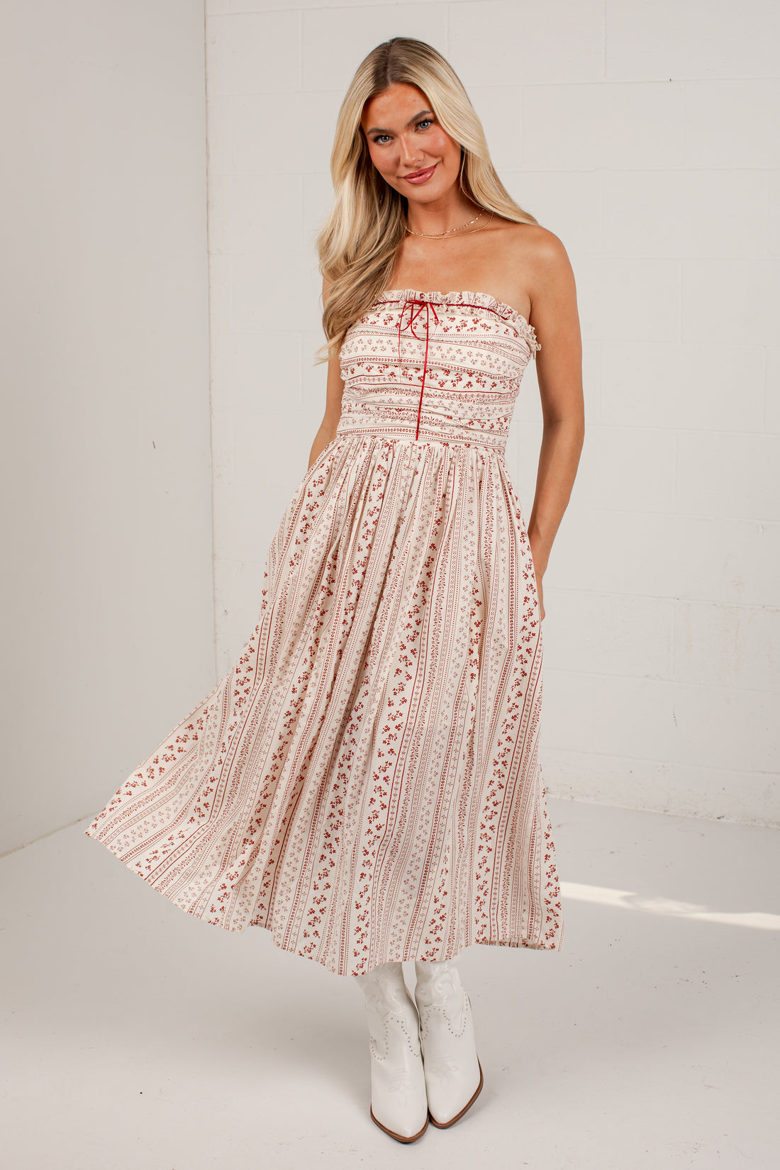 A woman modeling a strapless midi dress with a red and beige bohemian-style geometric pattern, paired with white cowboy boots, set against a white studio background.