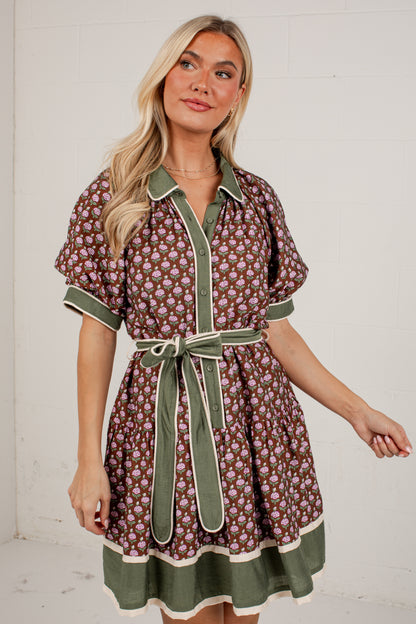 A woman modeling a short-sleeve button-front dress with an allover floral print in brown, green, and cream, featuring solid green trim, a matching green tie belt, and a green ruffled hem, set against a white brick wall.
