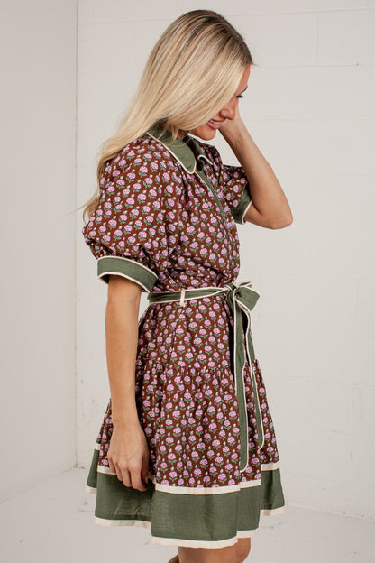 A woman wearing a short-sleeve button-front dress with an allover floral print in brown, pink, and green, featuring solid green trim and a matching green tie belt, standing against a white brick wall.